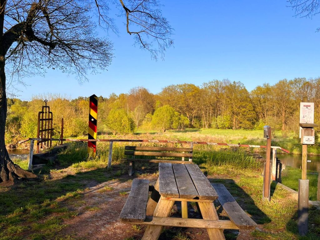 Ein Holztisch mit integrierten Bänken steht draußen im Sonnenschein. Im Hintergrund sind Bäume und ein deutscher Grenzpfosten zu sehen.